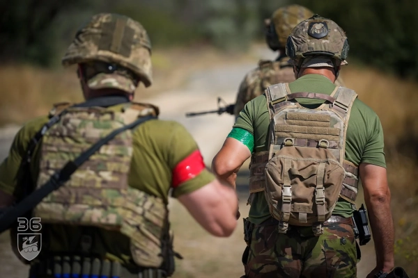 Троє військових у шоломах і захисному спорядженні рухаються ґрунтовою дорогою. На їхніх руках закріплені червоні та зелені стрічки.